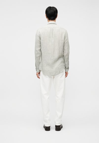 Άνδρας που στέκεται με την πλάτη γυρισμένη, φοράει μακρυμάνικο πουκάμισο με πράσινες και λευκές ρίγες, λευκό παντελόνι και σκούρα καφέ παπούτσια, πάνω σε λευκό φόντο.