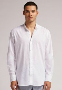 Camisa de lino blanca de manga larga con cuello mao, que presenta un cierre frontal de botones y una textura suave y ligera.