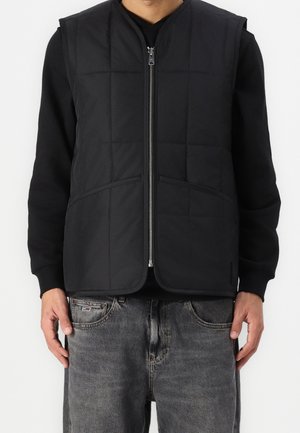 Mand iført sort quiltet vest med lynlås over sort langærmet skjorte og falmede grå jeans står mod en hvid baggrund.