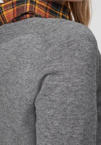 Nahaufnahme einer grauen Strickpullover-Schulter, die über einem rot, gelb und schwarz karierten Hemdkragen getragen wird, mit sichtbarem langen Haar.