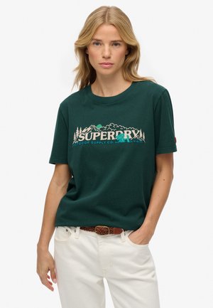 Kobieta ubrana w ciemnozieloną koszulkę Superdry z grafiką przyrody i białe spodnie, stojąca z ręką w kieszeni na białym tle.