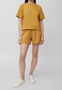 Camiseta de algodón amarilla mostaza con mangas cortas y cuello redondo, combinada con pantalones cortos a juego que cuentan con una cintura elástica con cordón. Zapatillas blancas.