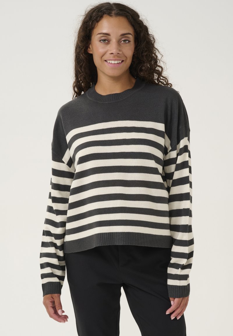Grau- und cremefarbener gestreifter Pullover mit langen Ärmeln und geripptem Ausschnitt, lockere Passform, kombiniert mit schwarzen Hosen. Glatte Stricktextur.