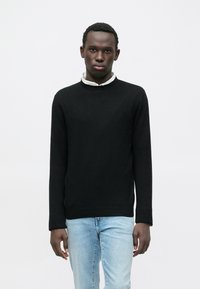 Suéter de punto negro con cuello redondo y mangas largas, usado sobre una camisa blanca con cuello, combinado con jeans azul claro.