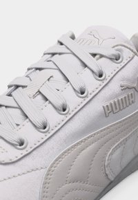 Gros plan sur une basket blanche Puma présentant des lacets, un logo cousu, et des détails en cuir texturé sur le côté et la languette.