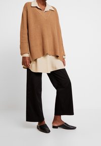 Beige ribbad stickad tröja med krage, lagd över en ljusbeige skjorta med knappar, tillsammans med svarta vida byxor och svarta slip-on skor.