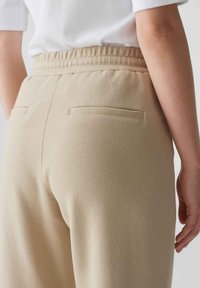 Beige, gestructureerde broeken met een aangesloten taille en elastische band. Voorzien van twee achterzakken met vierkante kleppen en subtiele stiksels.