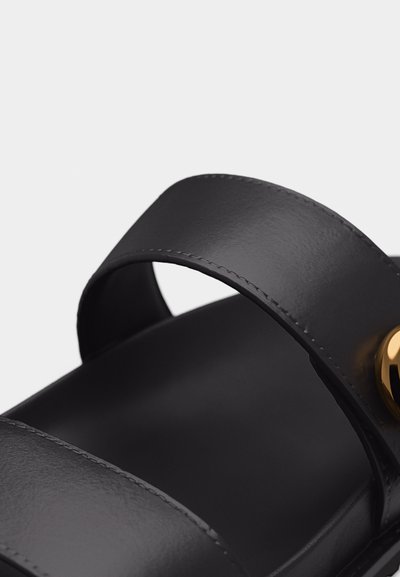 Μαύρο δερμάτινο λουράκι σανδάλι με εμφανή ραφή και χρυσή κυκλική αγκράφα πάνω σε μονόχρωμο φόντο.