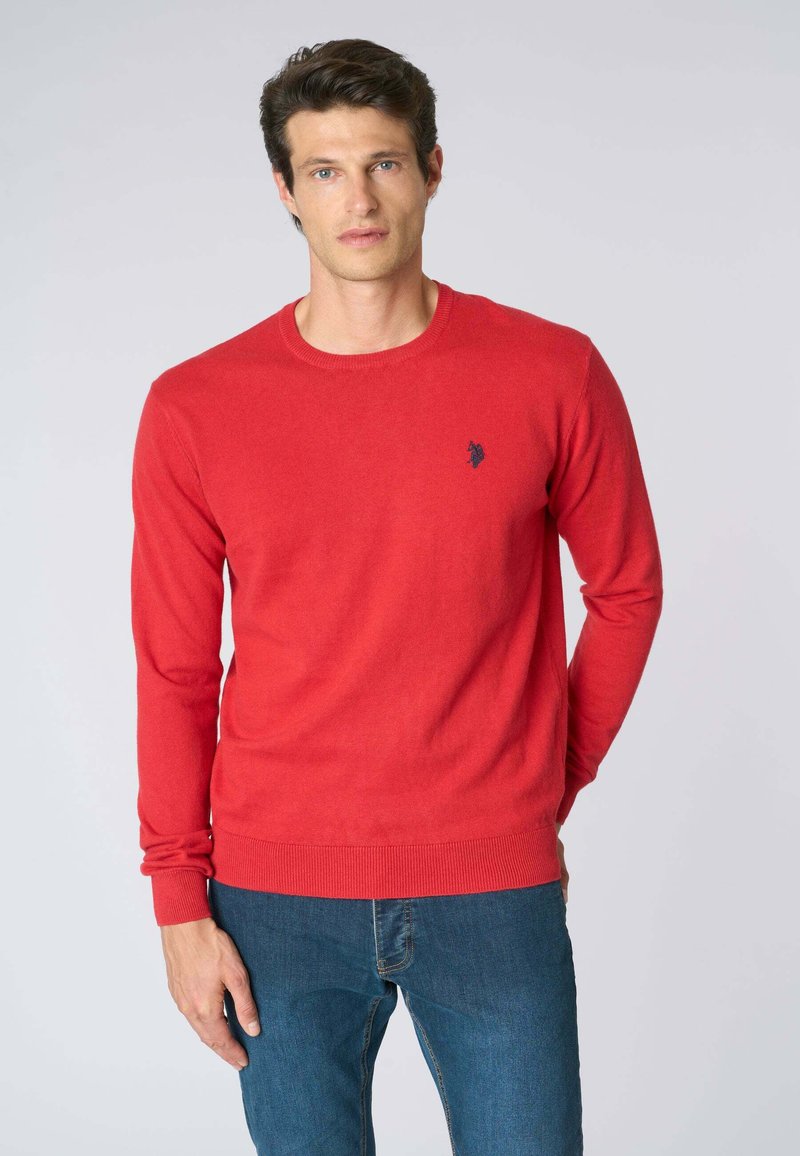 Maglione rosso lavorato a maglia con collo rotondo. Maniche e orlo a coste. Piccolo logo blu navy sul lato sinistro del petto. Indossato con jeans blu.