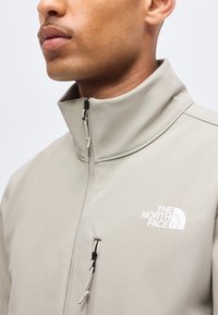 Gros plan d’un homme portant une veste zippée beige clair avec le logo "The North Face" sur la poitrine et un col montant.