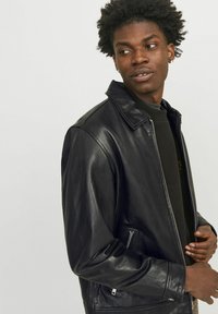 Black leather jacket with a zip front, collar, and zipper pockets. Soft texture and relaxed fit. Worn over a dark sweatshirt.