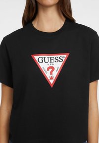 T-shirt en coton noir à manches courtes, présentant un grand logo triangulaire rouge et blanc avec le texte "GUESS U.S.A." et un design de point d'interrogation.