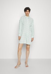 Vestido de camisa xadrez verde e branco com colarinho, mangas longas e fecho com botões. Possui um corte descontraído e cai a meio da coxa.