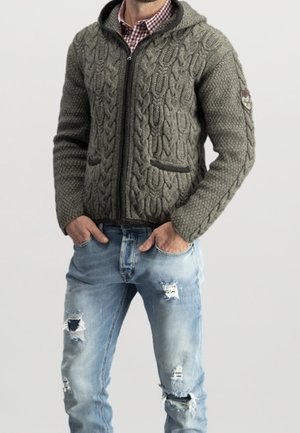 Homme portant un sweat à capuche gris en maille avec fermeture éclair et poches, par-dessus une chemise rouge à carreaux, et un jean bleu clair déchiré avec les mains dans les poches.