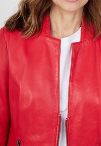 Červená kožená bunda na zip, s vysokým límcem a hladkým texture, kombinovaná s bílou košilí pod ní.