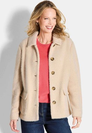 Veste en polaire texturée beige avec un col, fermeture à boutons à l'avant, poches latérales et coupe décontractée, portée sur un pull corail.