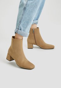 Bottes chelsea en suède beige avec un bout carré et un talon bloc. Fermeture éclair sur le côté, texture lisse, et jeans à revers visibles sur l'image.