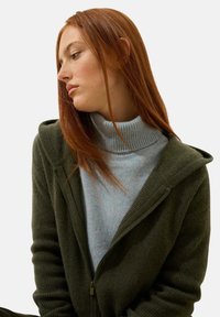 Haut à capuche zippé vert olive avec une texture côtelée, associé à un col montant côtelé bleu clair. Les deux vêtements ont une coupe décontractée.