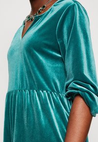 Tyrkysové sametové šaty s výstřihem do V a dlouhými nařasenými rukávy. Hrudník je dobře padnoucí, zatímco dolní část má nařasené detaily.