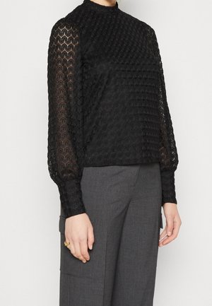 Persoon draagt een zwarte, getextureerde top met lange mouwen met pofmouwen en grijze, hooggesloten broek, hand ontspannen langs het lichaam.