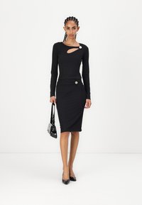 Robe noire à manches longues avec un décolleté découpé et un détail torsadé à la taille, ornée d'un bouton décoratif. Tissu lisse, silhouette ajustée.