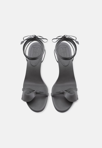 Μαύρα σανδάλια με ανοιχτά δάχτυλα, λεπτά λουριά στον αστράγαλο δεμένα σε φιόγκους και λεπτομέρεια από δεμένους δερμάτινους κόμπους πάνω από τα δάχτυλα, φωτογραφημένα από ψηλά.