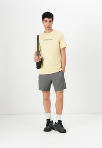 Camiseta amarilla de algodón con el logo de "Calvin Klein", pantalones cortos grises texturizados, calcetines blancos de caña corta y zapatillas deportivas negras.