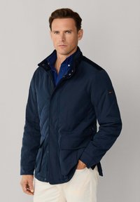 Marineblauwe doorgestikte jas met een getailleerd ontwerp, een hoge kraag, drukknoopsluiting en twee voorzakken; voorzien van een textuurstof en een contrasterende kraag.