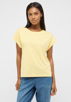 Woman with straight black hair wearing a light yellow short-sleeve top and blue jeans standing against a plain gray background.