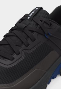 Μαύρο και σκούρο γκρι πεζοπορικό παπούτσι Columbia με μαύρα κορδόνια και μπλε λεπτομέρειες στη σόλα και στις θηλιές των κορδονιών.