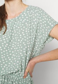 Femme portant une robe vert sauge à pois blancs irréguliers, la main posée sur la taille, avec des manches courtes et amples.