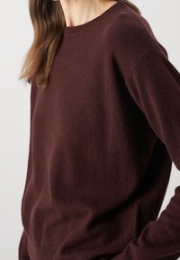 Femme portant un pull en tricot marron foncé à manches longues avec un col rond, sur un fond clair uni.