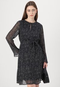 Čierne priesvitné šaty s dlhými rukávmi, majúce jemný bodkovaný vzor, detail v oblasti krku v tvare kľúča a viazaný pás. Stylizované s čiernou taškou.