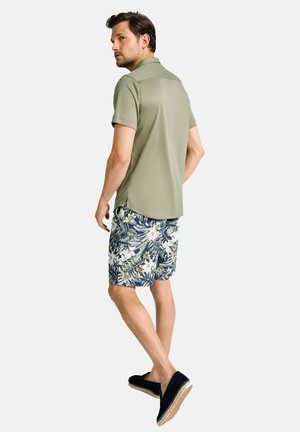 Olivgrünes Poloshirt mit kurzen Ärmeln, kombiniert mit blauen Badehosen mit Blumenmuster und weißen sowie gelben Akzenten, abgerundet durch dunkle Slipper.