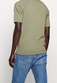 Olivově zelený svetr s krátkým rukávem a texturovaným povrchem, s žebrovanými manžetami a spodním lemem, v kombinaci se světle modrými džíny s kapsami na zadní straně.