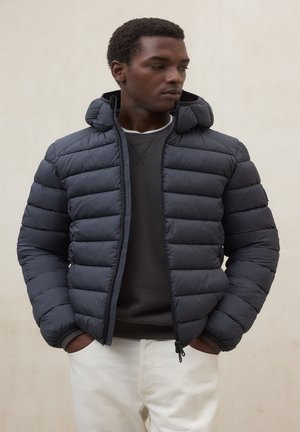 Uomo con le mani in tasca indossa una giacca a vento grigio scuro sopra una felpa nera e pantaloni bianchi, guardando verso sinistra su uno sfondo semplice.