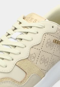 Λευκό και μπεζ αθλητικό παπούτσι με υφή δέρματος, χρυσές λεπτομέρειες και λογότυπο. Διαθέτει επίπεδα κορδόνια και σχεδιασμό σόλας σε αντίθετο χρώμα.