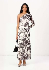 Robe maxi asymétrique en crème avec des taches abstraites noires, fabriquée à partir d'un tissu doux. Le modèle tient un petit sac noir et porte des chaussures grises.