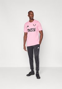 Pink t-shirt with black "Messi" logo, worn with black track pants featuring white stripes and black shoes. Smooth texture, sporty design.