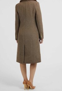 Cappotto in misto lana marrone con taglio dritto, polsini con bottoni e spacco posteriore. Abbinato a scarpe con tacco alte beige, che mostrano un design pulito.