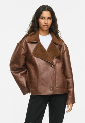 Femme aux longs cheveux foncés portant une veste en cuir retourné marron par-dessus une chemise blanche et un pantalon noir, debout devant un fond blanc uni.