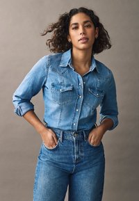 Jasnoniebieska koszula dżinsowa z długimi rękawami, zapinana na guziki, z dwoma kieszeniami na piersi, w połączeniu z dżinsami w wysokim stanie w kolorze niebieskim, na neutralnym tle.