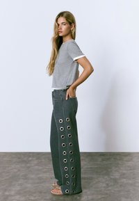 Mujer con largo cabello rubio que lleva una camiseta de manga corta gris y pantalones anchos oscuros con grandes ojales metálicos, de pie sobre un suelo de concreto.