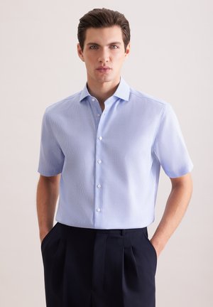 Giovane uomo che indossa una camicia azzurra a maniche corte infilata in pantaloni neri a vita alta con pieghe, con le mani nelle tasche.