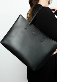 Zwarte leren tote bag met een gladde textuur, platte rechthoekige vorm, dubbele banden en een klein gouden logo accent aan de zijkant.