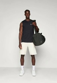 Camiseta sin mangas negra, pantalones cortos beige, sosteniendo un bolso de deporte negro. Calzado blanco, de pie sobre un fondo neutro. Diseño simple, ropa casual.