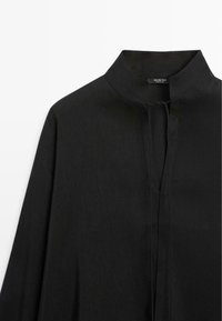 Schwarzes Hemd mit langen Ärmeln, Mandarin-Kragen und verdeckter Knopfleiste, auf weißem Hintergrund dargestellt.