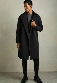 Trench coat negru, cu două rânduri de nasturi și reveruri late, cu detalii cu cataramă, asortat cu un top din tricot gri și pantaloni negri conici, pe un fundal neutru.