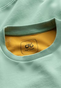 Collo della maglietta in cotone verde chiaro con texture a coste. L'etichetta interna presenta uno sfondo giallo con un logo nero e il testo "KAFT.COM".