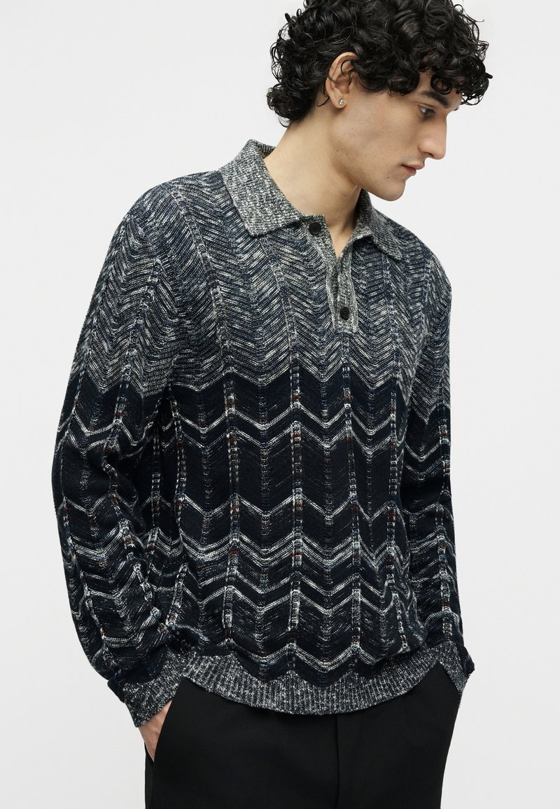 Young man with curly hair wearing a grey and black patterned knit sweater with a collar and black pants, standing against a plain background.